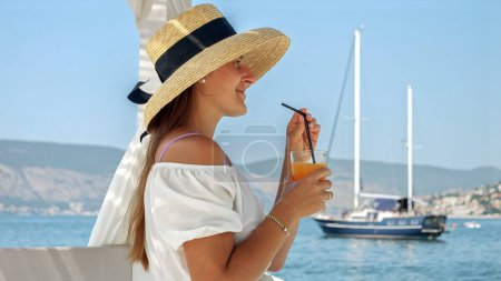 Téléchargez les photos : Belle femme portant un chapeau de paille, assise dans un kiosque sur la jetée de la mer en bois. Elle aime son jus d'orange ou son cocktail, regarder les yachts dans la mer, incarnant un voyage de luxe, le bonheur de l'été - en image libre de droit