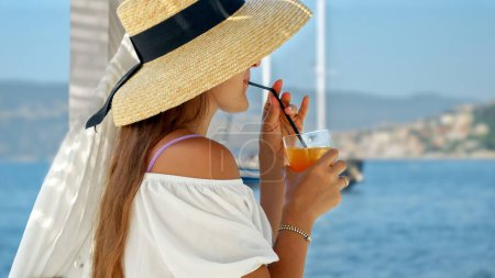 Téléchargez les photos : Femme dans un chapeau de paille se détend dans un belvédère sur une jetée de mer en bois. Sirotant son verre, elle admire les yachts en mer, incarnant le voyage de luxe et la joie de l'été - en image libre de droit