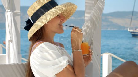 Foto de Mujer con sombrero de paja, en un cenador en un muelle de madera. Bebiendo su bebida, disfruta de yates en el mar, simbolizando viajes de lujo y verano - Imagen libre de derechos