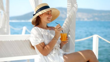 Foto de Mujer con sombrero de paja se instala en un mirador en un muelle de madera. Bebiendo su bebida, encuentra paz observando el mar, encarnando viajes de lujo y verano. - Imagen libre de derechos