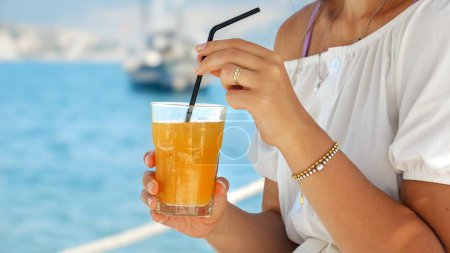 Téléchargez les photos : Gros plan de la femme assise dans le café de la plage de la mer et agitant le jus d'orange avec de la paille. Concept d'été, détente, profiter de l'océan - en image libre de droit