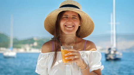 Téléchargez les photos : Portrait de femme souriante heureuse dans un grand chapeau profitant de son cocktail ou jus de fruits frais contre la mer et les yachts vue. - en image libre de droit