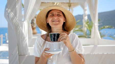 Foto de Retrato de una mujer sonriente con sombrero de paja bebiendo café en el café de playa en un día soleado y ventoso - Imagen libre de derechos
