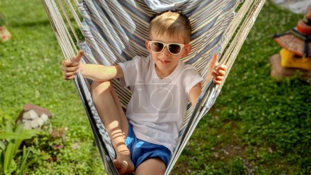 Téléchargez les photos : Dans un jardin, un garçon ravi portant des lunettes de soleil profite de la balançoire d'un hamac, savourant l'essence de l'été, les joies simples de l'enfance, la tranquillité et la magie des vacances d'été. - en image libre de droit
