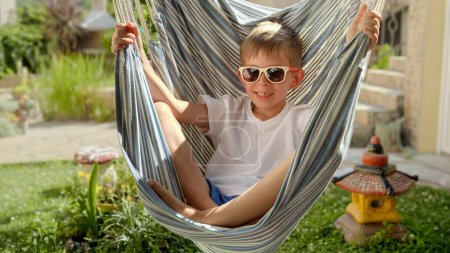 Foto de Un muchacho joven sonriente se relaja en el balanceo suave de la hamaca en un jardín, encapsulando las sensaciones del verano, de los recuerdos felices de la infancia, del ocio, y de los descansos preciados del verano. - Imagen libre de derechos