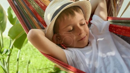 Téléchargez les photos : Un garçon joyeux dans le chapeau se détend dans un hamac de jardin, capturant l'essence de l'été, enfance heureuse, et bonheur de vacances. - en image libre de droit