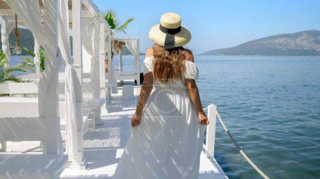 Photo for Young woman in long light dress and straw hat walking on the wooden pier at sea beach. Concept of holiday summertime, vacation, travel, journey, relaxing at ocean. - Royalty Free Image
