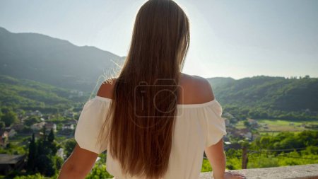 Téléchargez les photos : Vue arrière de la femme brune jouissant d'une vue sur l'allée et les montagnes avec la lumière du coucher du soleil depuis sa terrasse de la villa. - en image libre de droit