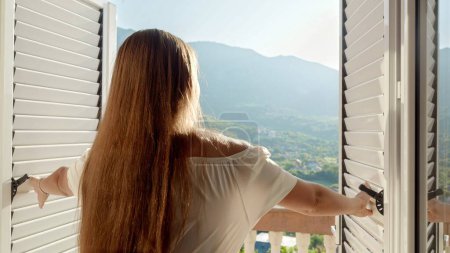 Téléchargez les photos : La gracieuse jeune brune ouvre les portes de son balcon, se promène sur la terrasse et admire le lever ou le coucher du soleil de montagne. Essence des vacances d'été. - en image libre de droit
