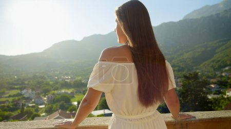 Téléchargez les photos : Une jeune femme brune, vêtue d'une robe fluide, se tient sur la terrasse ou le balcon de la villa, profitant du coucher de soleil sur la montagne. Été, bonheur de la montagne, sérénité des vacances et voyages. - en image libre de droit