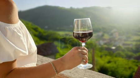 Foto de Primer plano de la joven morena sosteniendo una copa de vino tinto contra el atardecer y la vista a la montaña desde el balcón o terraza de la villa. Relajación femenina, concepto de viaje, verano y vacaciones - Imagen libre de derechos