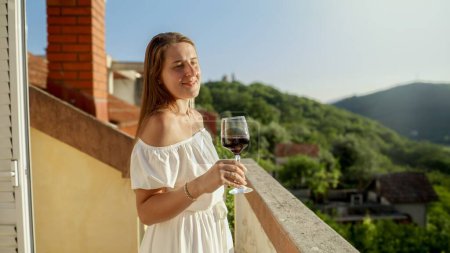 Téléchargez les photos : Jeune femme brune souriante, tenant gracieusement un verre de vin rouge, sur fond de coucher de soleil envoûtant et de vue sur la montagne depuis le balcon ou la terrasse de la villa - en image libre de droit