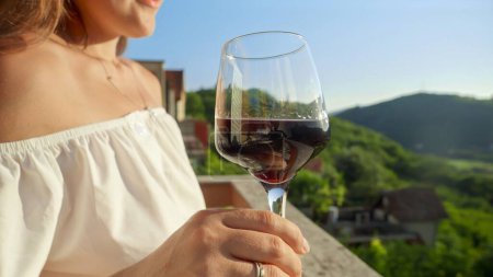 Photo for Young woman with glass of red wine gracefully held, merges with the magic of a mesmerizing sunset and mountain view from the villa balcony or terrace. Travel, summertime, and holiday serenity. - Royalty Free Image