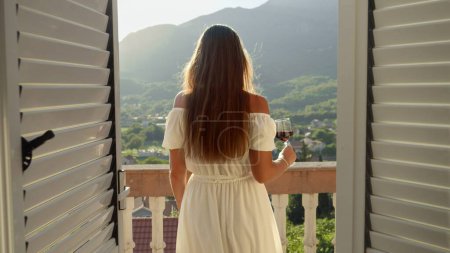 Téléchargez les photos : Femme brune élégante tenant un verre de vin rouge marchant sur le balcon ou la terrasse de la villa et regardant le coucher du soleil sur les montagnes. - en image libre de droit