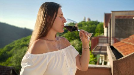 Téléchargez les photos : Gros plan d'une élégante brune buvant du vin rouge dans un verre debout sur un balcon ou une terrasse de la villa en montagne. Profiter du vin, célébration des vacances, vacances d'été - en image libre de droit