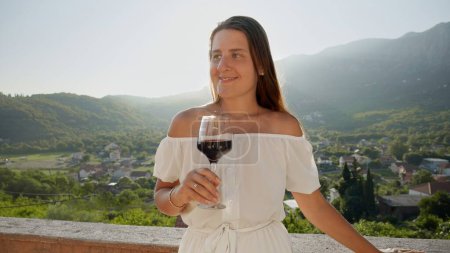 Téléchargez les photos : Portrait de femme brune souriante tenant un verre de vin rouge et souriant sur le balcon de la villa en montagne contre le coucher du soleil - en image libre de droit
