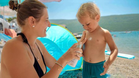 Photo for Portrait of smiling loving mother applying sunscreen lotion on her little toddler relaxing on sea beach. - Royalty Free Image