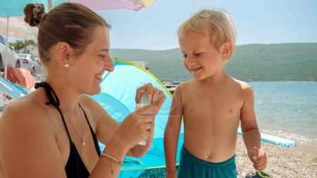 Téléchargez les photos : Jeune mère appliquant de la crème solaire sur son bébé fils tout en se relaxant sur la plage de la mer par une journée ensoleillée. Concept de santé, vacances d'été, vacances avec les enfants. - en image libre de droit