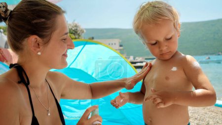 Photo for Closeup of mother and baby son using sunscreen lotion while having time on sea beach. - Royalty Free Image