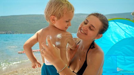 Photo for Young mother ensuring her baby son is protected with sunscreen spray from the sun's rays as they enjoy a sunny day at the beach. Healthcare during summer vacations. - Royalty Free Image