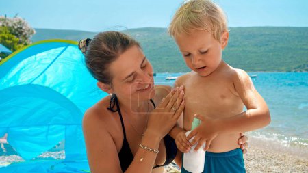 Téléchargez les photos : Aimant jeune mère appliquant diligemment un spray solaire à son fils sur une plage ensoleillée. Concept de soins de santé et vacances en famille. - en image libre de droit