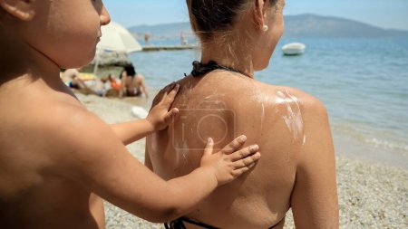 Téléchargez les photos : Gros plan de petit garçon frottant la crème solaire UV dans le dos de sa mère avant de se détendre sur la plage ensoleillée de la mer. - en image libre de droit