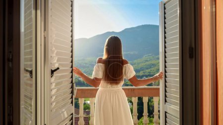 Photo for Young brunette woman in a flowing dress opens balcony doors, steps onto the terrace, and revels in a breathtaking mountain sunrise or sunset. Essence of summertime holiday, vacation, nature appreciation, and travel. - Royalty Free Image