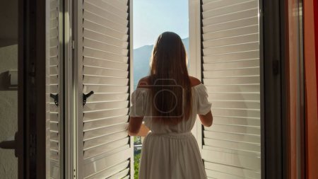 Junge brünette Frau in langem Kleid öffnet Balkontüren, geht auf die Terrasse und genießt Sonnenaufgang oder Sonnenuntergang über den Bergen. Sommerurlaub, Urlaub, Natur genießen und Reisen.