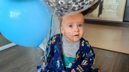 Foto de Feliz niño sonriente sentado en el suelo y jugando con globos de aire de colores en su primera fiesta de cumpleaños. - Imagen libre de derechos