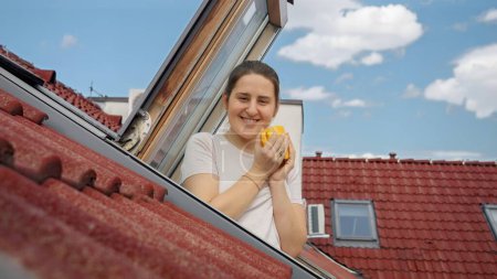 Foto de Mujer con una cálida sonrisa saborea su té o café junto a una ventana del ático, disfrutando del encantador paisaje urbano de tejados de tejas rojas en una antigua ciudad europea. - Imagen libre de derechos