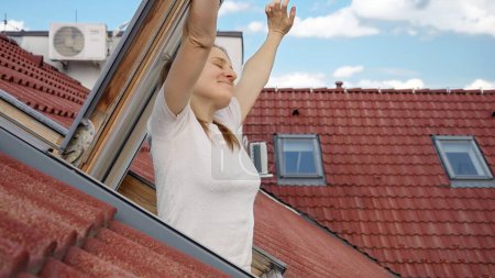 Téléchargez les photos : Femme brune souriante ouvre la fenêtre du grenier sous le toit et étire les mains. - en image libre de droit
