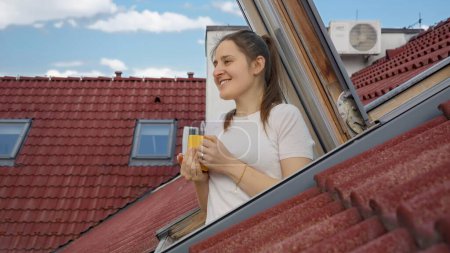 Téléchargez les photos : Heureuse femme souriante regarde à travers la fenêtre ouverte grenier et boit du thé ou du café à partir d'une tasse le matin - en image libre de droit