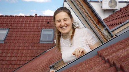 Téléchargez les photos : Heureuse femme souriante et riante regardant par la fenêtre ouverte du grenier dans son appartement sous le toit carrelé rouge. - en image libre de droit