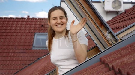 Téléchargez les photos : Heureuse femme souriante regardant par la fenêtre du grenier et agitant la main dans la caméra. - en image libre de droit