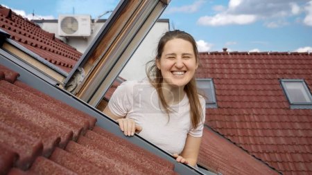 Téléchargez les photos : Heureuse femme souriante et riante regardant par la fenêtre ouverte grenier et vagues main dans la caméra. - en image libre de droit