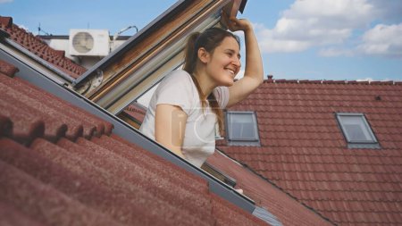 Téléchargez les photos : Heureuse femme brune qui regarde par la fenêtre d'un grenier, accentuée par la présence d'un toit traditionnel en tuiles rouges. - en image libre de droit