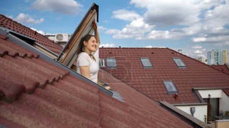 Téléchargez les photos : Heureuse femme brune souriante ouvrant la fenêtre du grenier et respirant de l'air frais tout en regardant le paysage urbain avec des toits carrelés rouges. - en image libre de droit