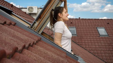 Téléchargez les photos : Femme souriante en regardant par la fenêtre d'un grenier sous un toit carrelé rouge et respirant l'air frais. - en image libre de droit