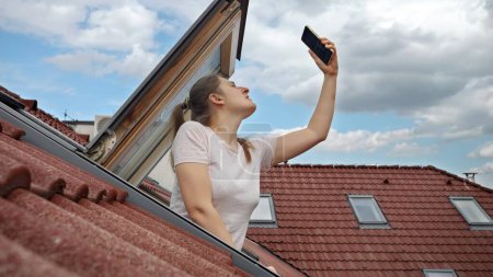 Téléchargez les photos : Jeune femme rencontre des problèmes de signal avec GPS ou 5G sur son smartphone tout en regardant par une fenêtre ouverte grenier, déterminé à restaurer la connectivité. - en image libre de droit