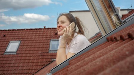 Téléchargez les photos : Femme souriante parlant sur son téléphone tout en regardant par une fenêtre ouverte du grenier, en admirant la vue pittoresque des toits de tuiles rouges dans une vieille ville européenne - en image libre de droit