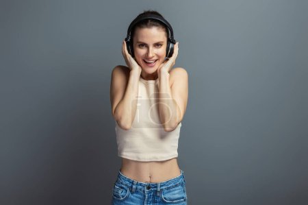 Foto de Joven mujer alegre disfrutando de la música con auriculares, vistiendo top casual, aislado sobre fondo gris estudio - Imagen libre de derechos