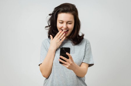 Foto de Hermosa chica con un teléfono en la mano se sorprende por algo que ve en la pantalla, aislado en el fondo gris. - Imagen libre de derechos