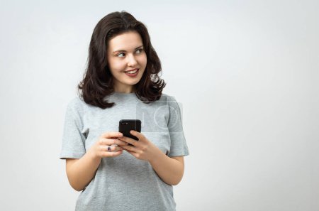 Foto de Alegre hermosa joven sosteniendo un teléfono celular y mirando hacia otro lado, espacio de copia, aislado sobre fondo gris - Imagen libre de derechos