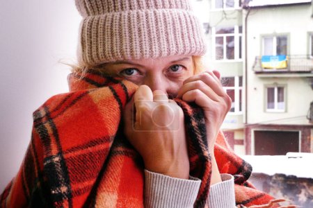 Photo for Woman sits in apartment near window in winter hat and wrapped in a blanket. Increase price of natural gas for heating in Ukraine. Energy crisis in Ukraine. Ukrainian suffer from cold in winter and freeze in their homes - Royalty Free Image