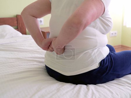 Photo for Over weight blond middle-aged woman suffering from back ache on the bed, healthcare and problem, Woman rubbing aching back, indoors - Royalty Free Image