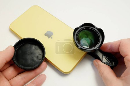 Foto de Dnipro Ukraine - 04 04 22: yellow smartphone iPhone with external Apexel lens on clip on white. An additional portrait lens and clip are supplied with the mobile phone, closeup - Imagen libre de derechos