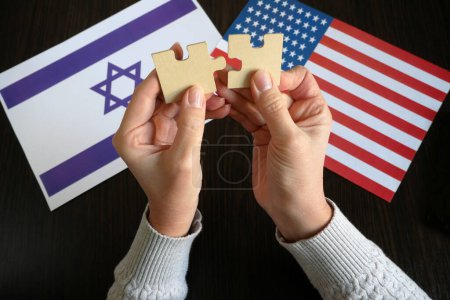 Téléchargez les photos : Mains tenir deux pièces d'un puzzle sur le fond des drapeaux Etats-Unis et Israël, amitié et partenariat de deux pays concept, gros plan - en image libre de droit