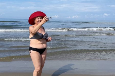 Senior woman wearing in swimsuit staying on the sea beach and speaking with mobile phone in sunny day. Mature woman smiles holding cellphone in hand. Concept of roaming, wifi, vacation, freedom and happy retirement. Outdoors