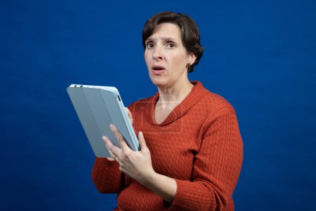 Foto de Una mujer reacciona a su lector electrónico con un suéter naranja de clima frío sobre un fondo azul mirando a la cámara. - Imagen libre de derechos