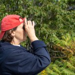Middle-aged woman looking through binoculars wearing a bright red baseball cap on a sunny day.